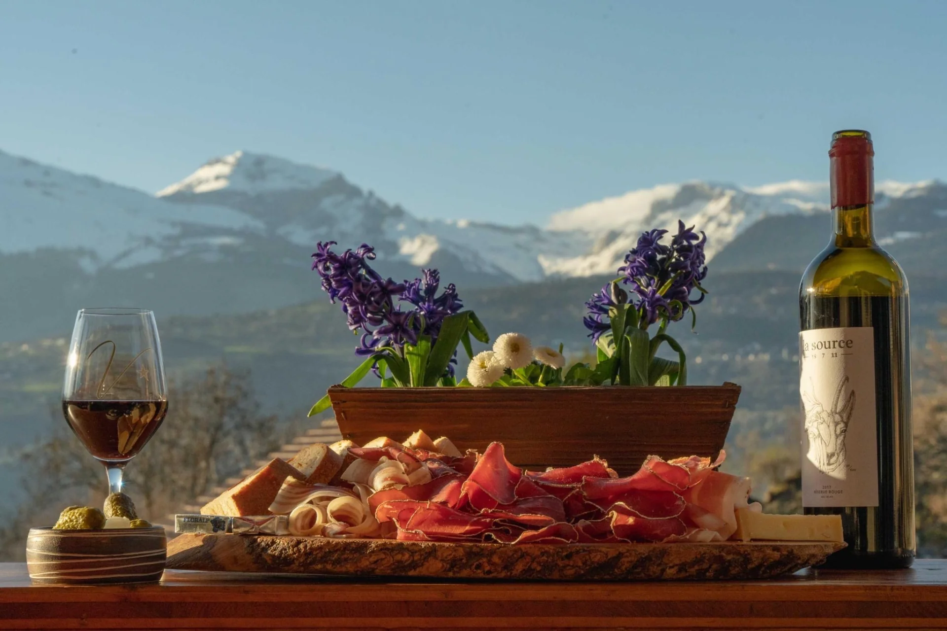 Assiette valaisanne avec viande séchée, verre de vin et fleurs, les Alpes en arrière-plan